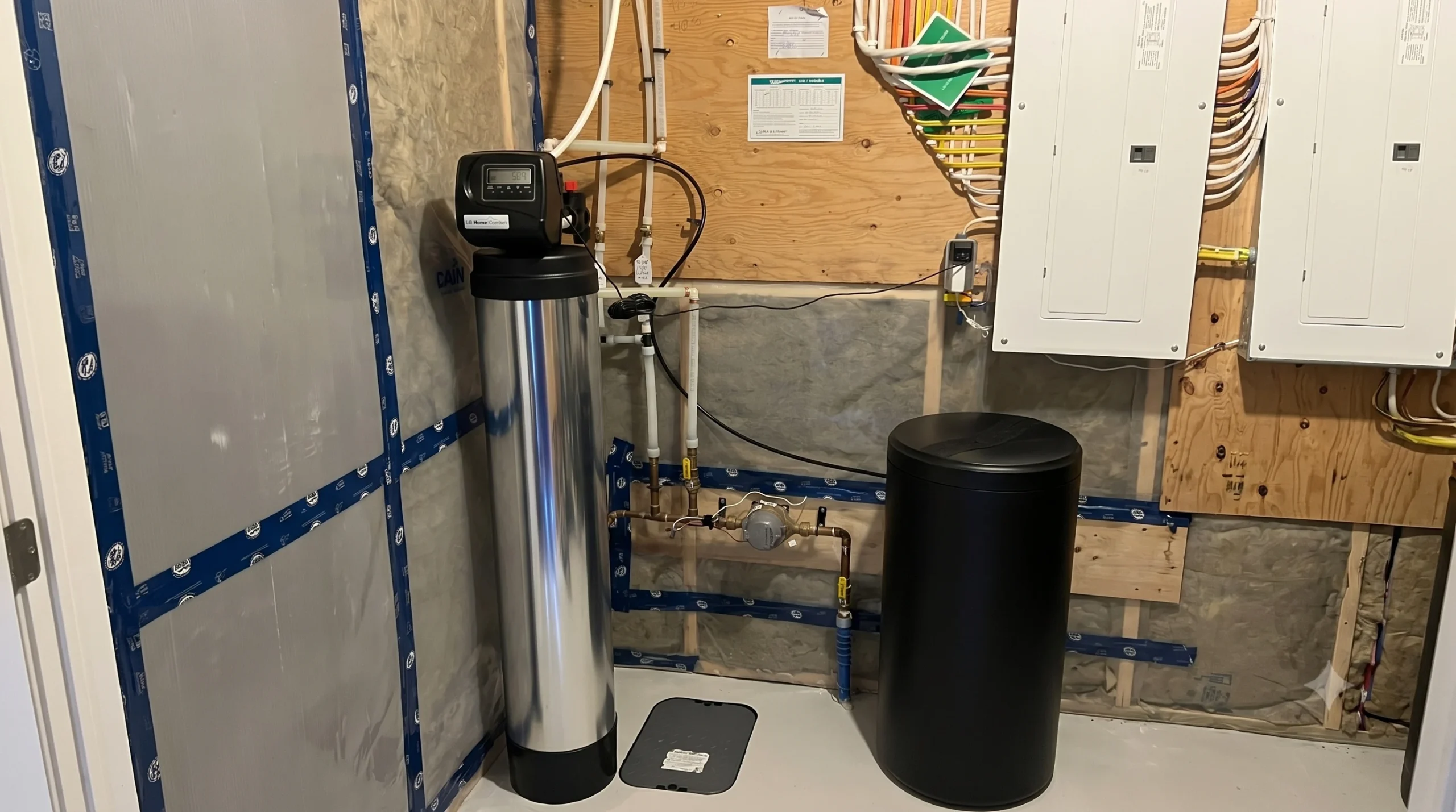 Newly installed dual-tank ion-exchange water conditioning system connected to residential PEX piping alongside utility room electrical distribution panels.