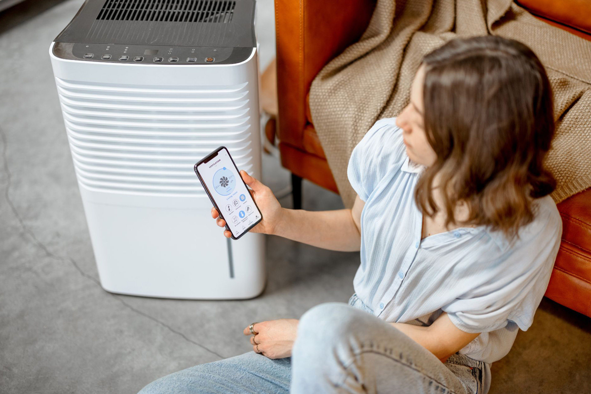 A woman sits near an air purifier and uses her phone to control it