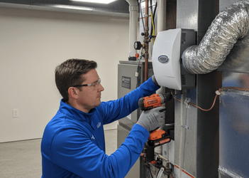 Gloved expert fastening white AprilAire evaporative humidifier module onto galvanized HVAC airflow ductwork utilizing orange cordless power drill inside illuminated residential mechanical basement