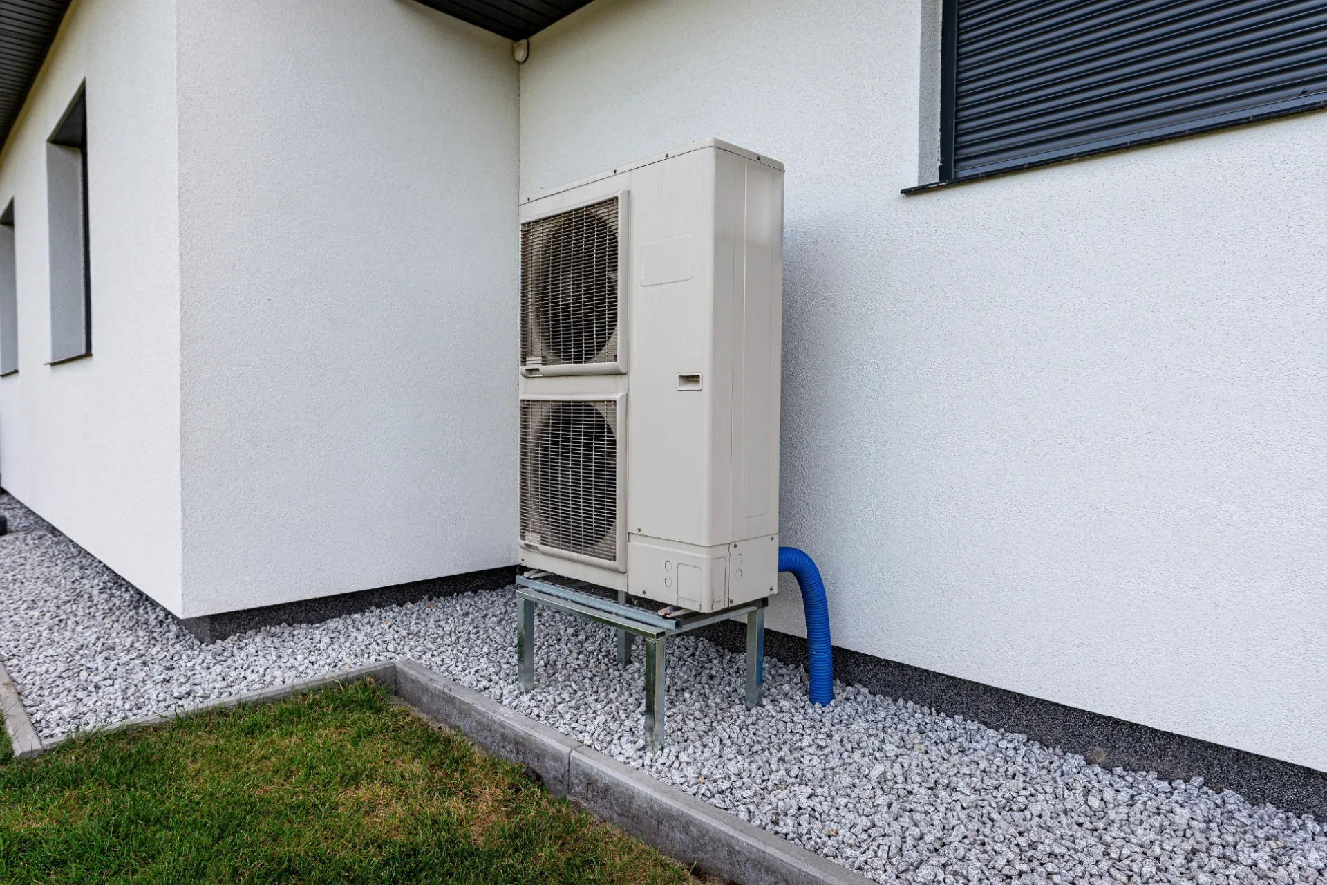 Outdoor heat pump unit mounted on a metal stand beside a white house, set on gravel with a blue conduit running into the wall